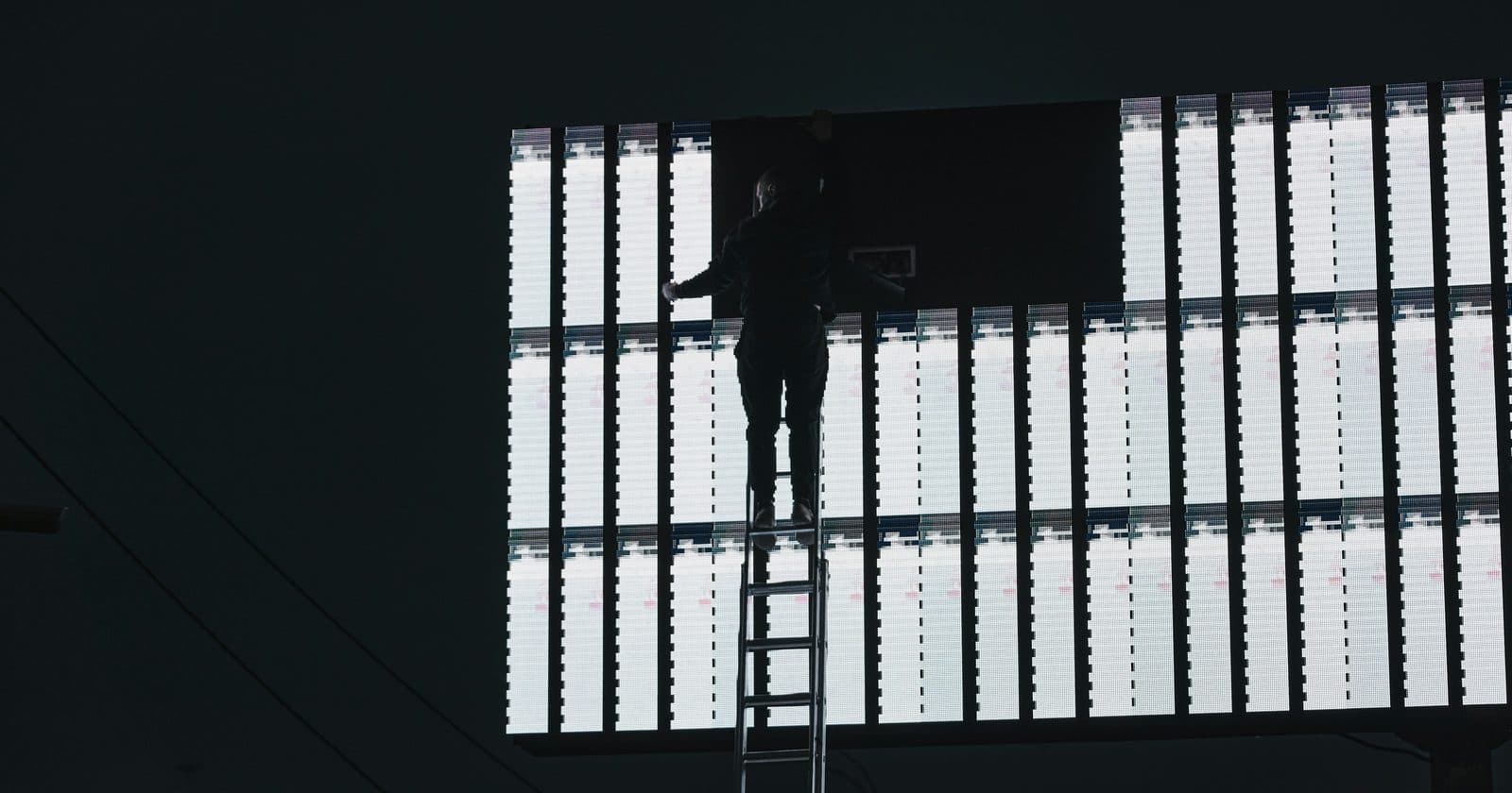 A technician on a ladder working on a large partly-lit LED billboard at night